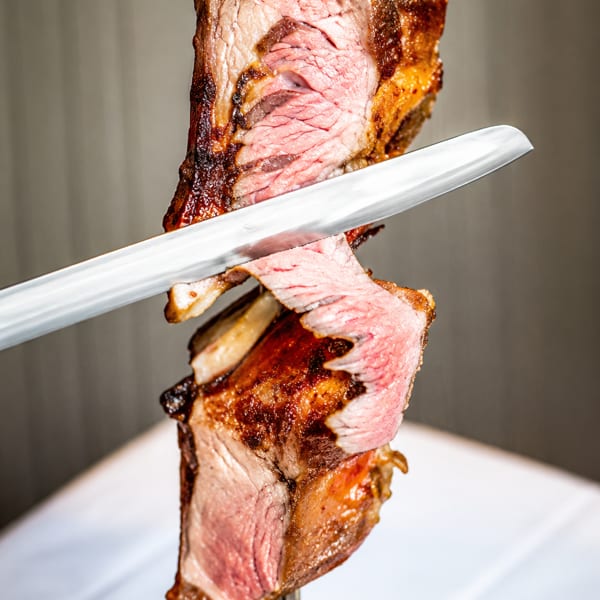 A chef carves a slice of perfectly cooked, medium-rare prime rib from a rotisserie skewer.
