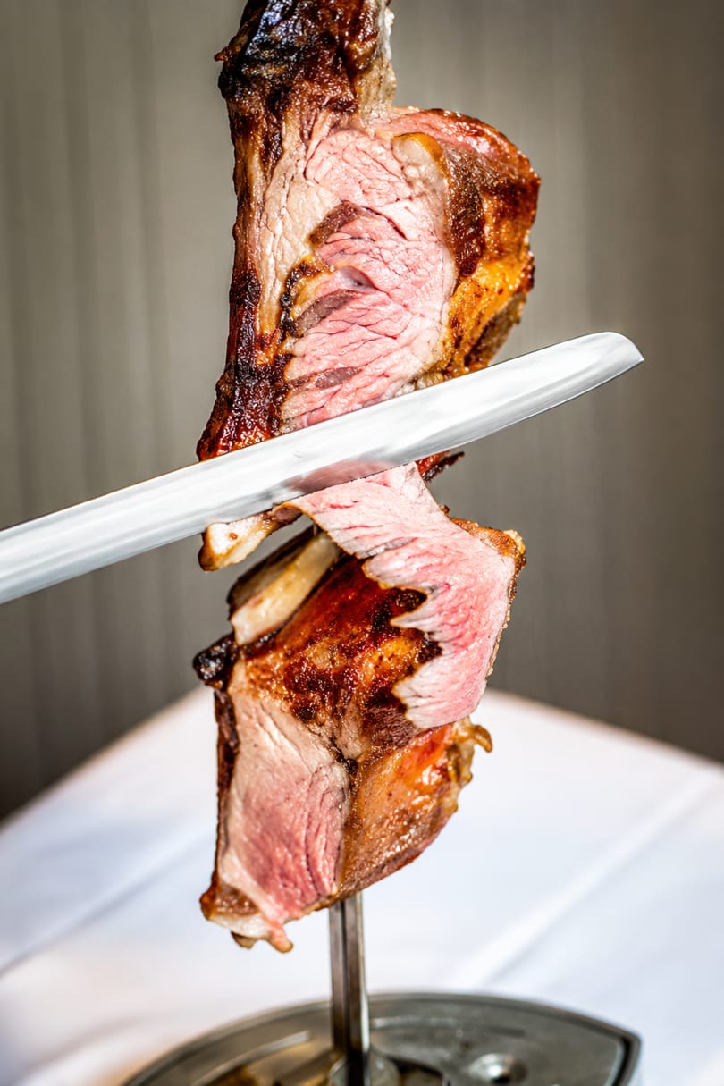 A chef carves a slice of perfectly cooked, medium-rare prime rib from a rotisserie skewer.