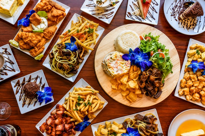 A wooden table laden with various dishes, including fried chicken, fries, pasta, rice, and desserts.