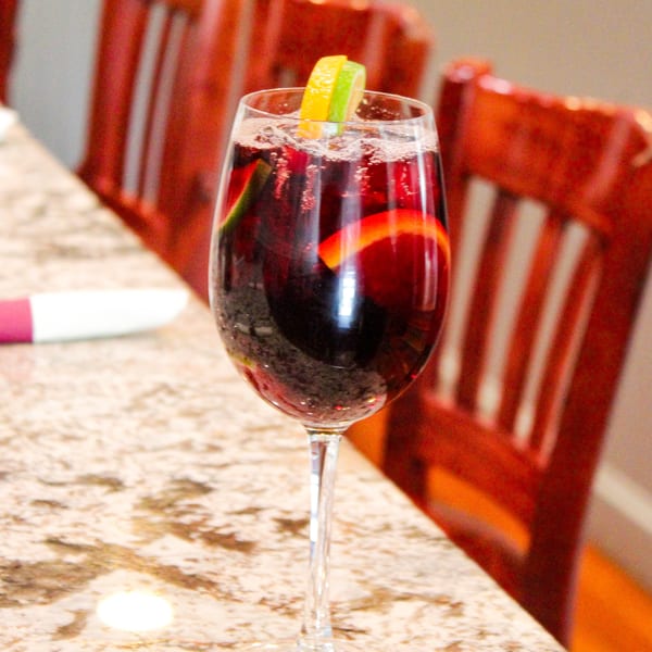 A glass of red sangria with orange and lime slices sits on a speckled countertop.