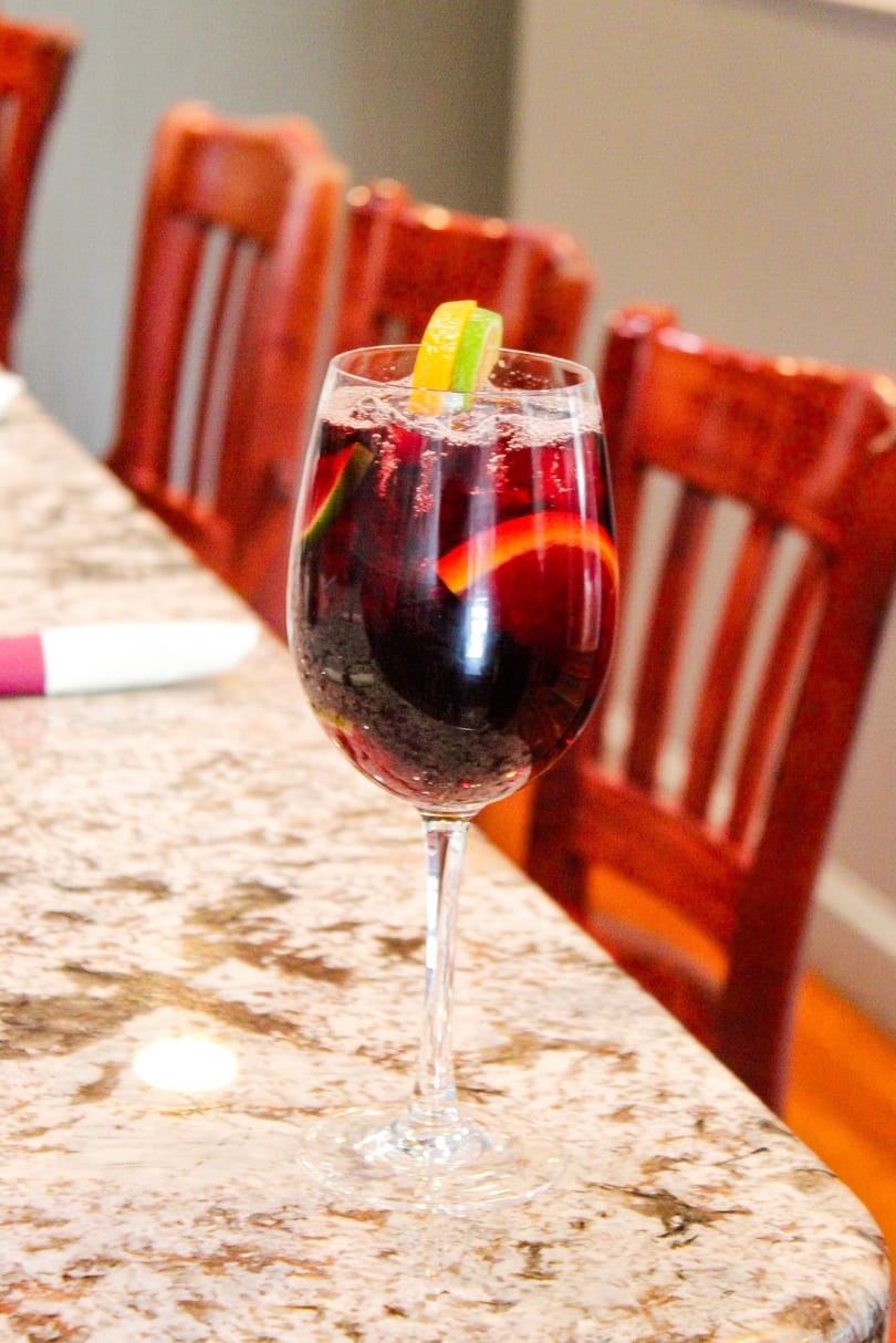A glass of red sangria with orange and lime slices sits on a speckled countertop.