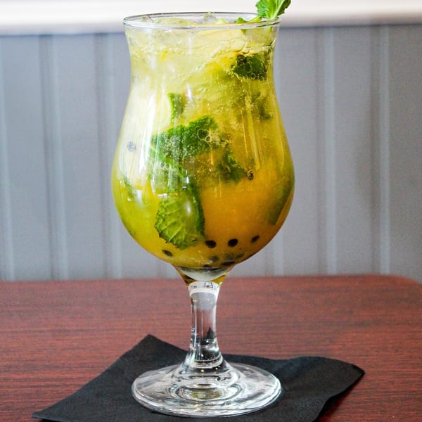 A vibrant yellow passion fruit mojito with mint leaves and seeds in a hurricane glass on a dark table.
