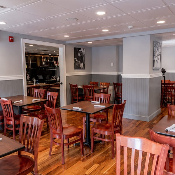 Empty dining room with wooden tables and chairs, a bar area visible through an open doorway.