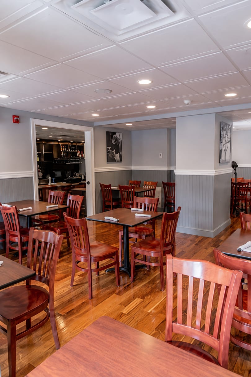 Empty dining room with wooden tables and chairs, a bar area visible through an open doorway.