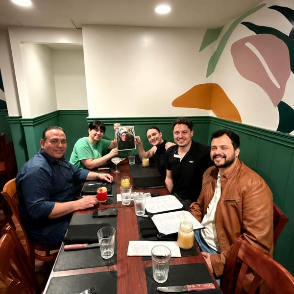 Five men smiling at a restaurant table with drinks and menus, one holding up a photo.