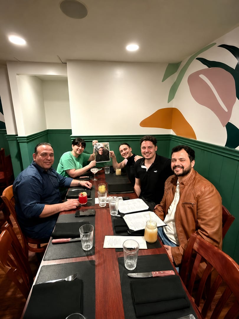 Five men smiling at a restaurant table with drinks and menus, one holding up a photo.