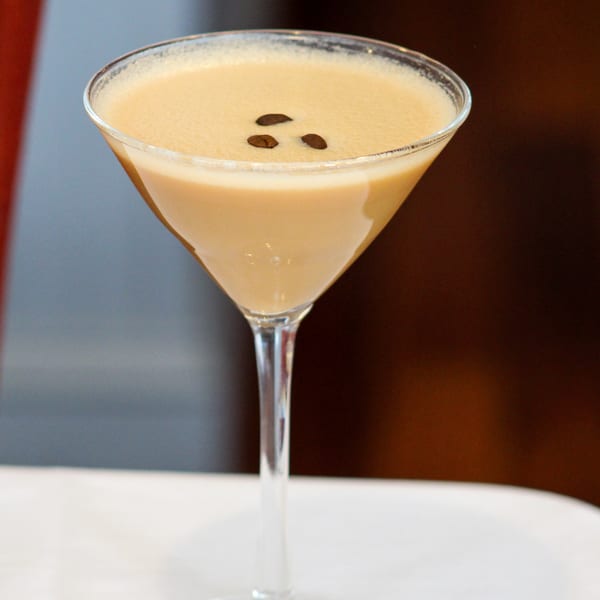 A creamy espresso martini in a stemmed glass, garnished with three coffee beans on a white tablecloth.