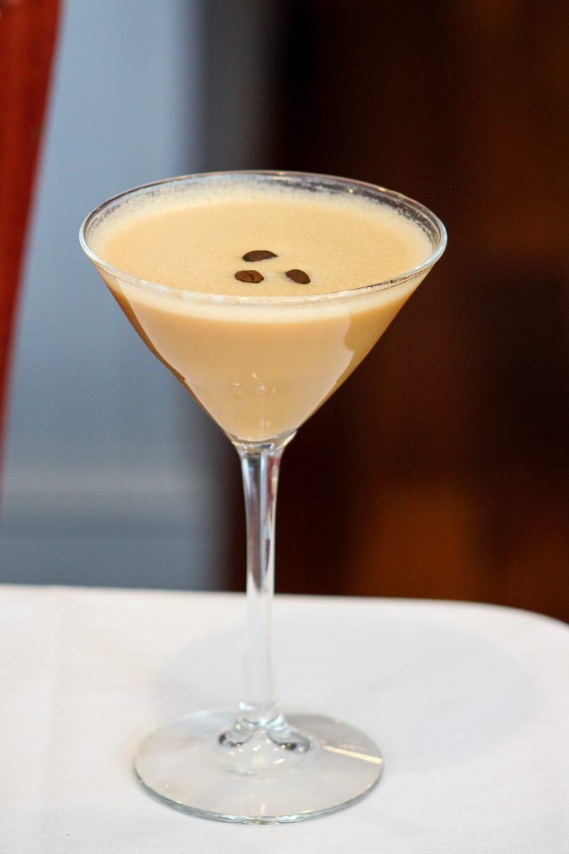 Espresso martini in a stemmed glass, garnished with three coffee beans, on a white tablecloth.