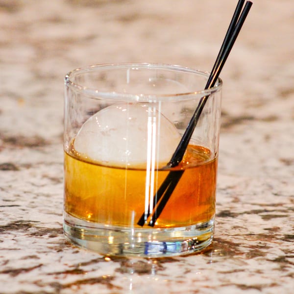 Amber cocktail with a large spherical ice cube and two black straws on a speckled countertop.