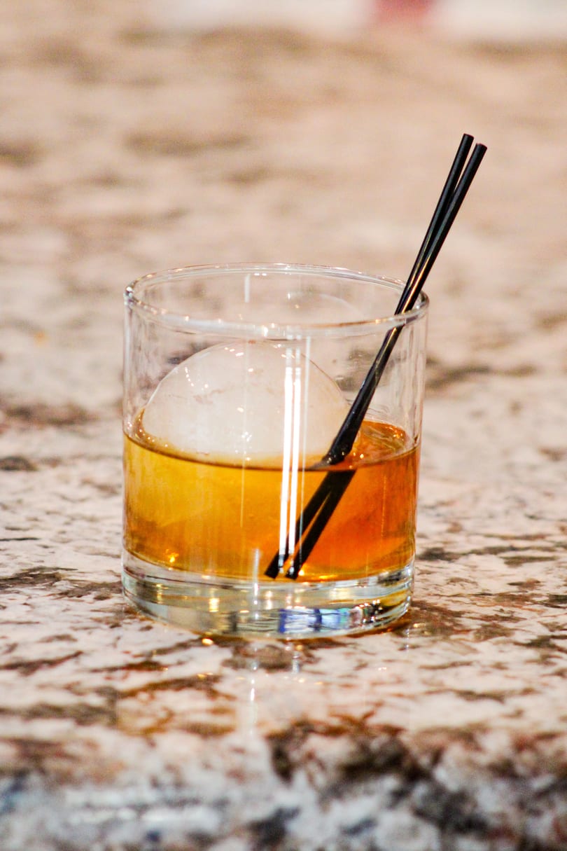 Amber cocktail with a large spherical ice cube and two black straws on a speckled countertop.