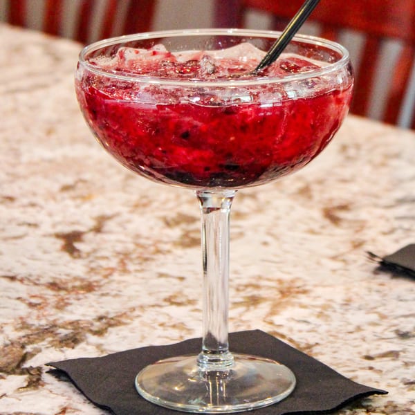 A vibrant red berry cocktail with ice and a black straw, on a marble table with red chairs.