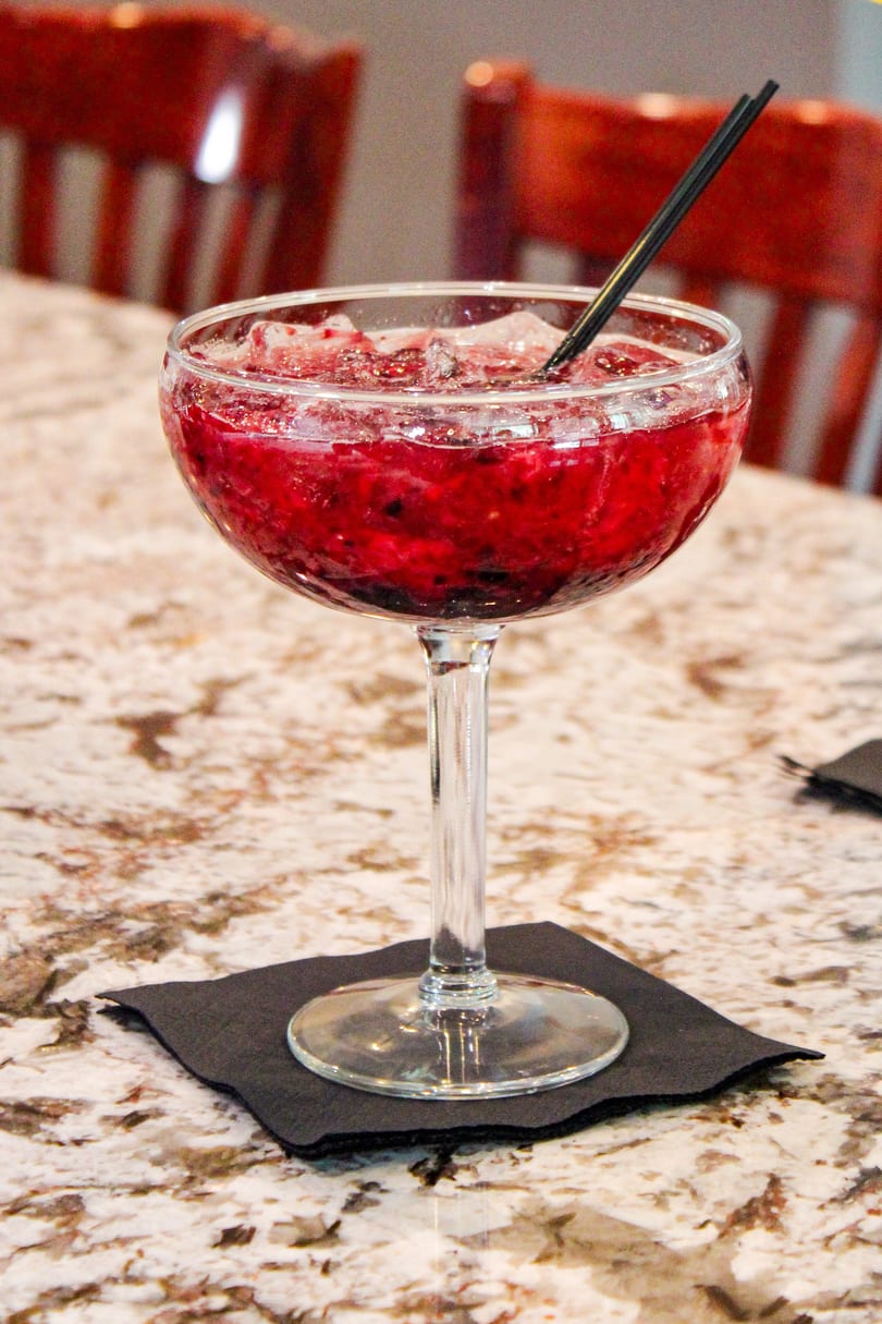 A vibrant red berry cocktail with ice and a black straw, on a marble table with red chairs.